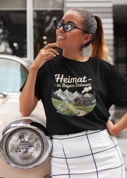 Junge Frau, an Auto gelehnt, mit Sonnenbrille. Sie trägt schwarzes T-Shirt mit Motiv „Heimat – In Bayern dahoam“, inklusive Berghintergrund und Almhütte, Oberkörperansicht