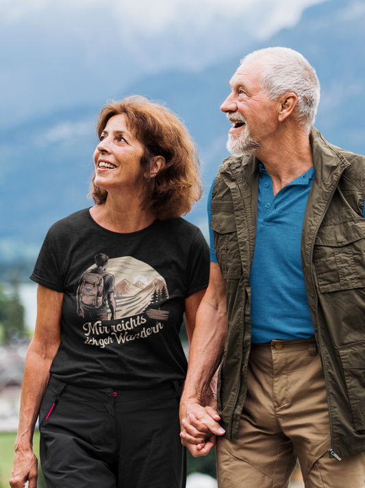 Älteres Paar, Hand in Hand, lachend. Die Frau trägt ein schwarzes Shirt mit Print und eine schwarze Wanderhose. Der Mann trägt eine offene Weste und darunter ein blaues Shirt.