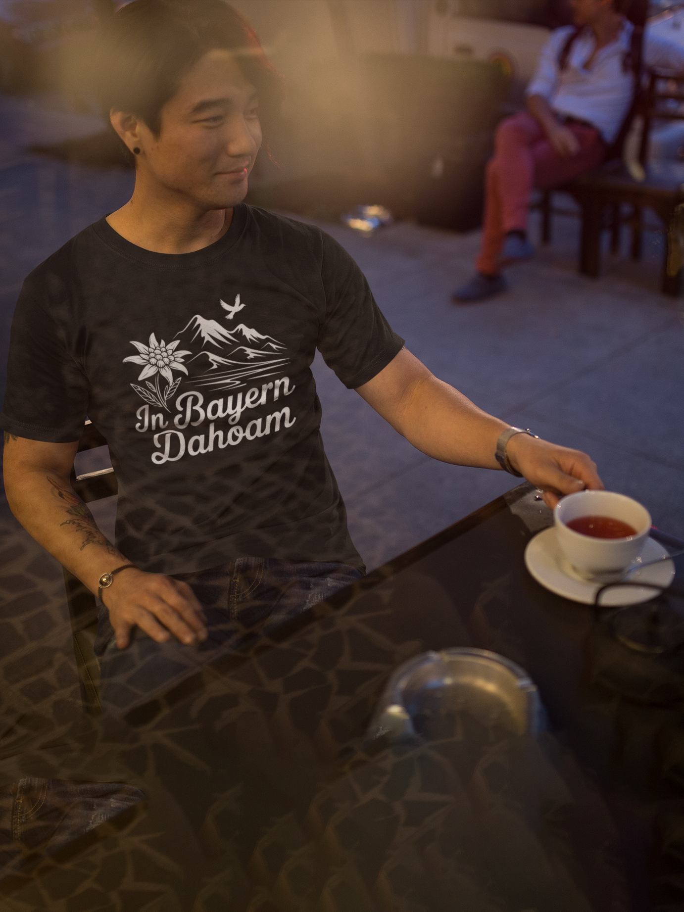Junger asiatischer Mann trägt schwarzes T-Shirt mit Bergpanorama und „In Bayern dahoam“-Schriftzug. Er sitzt in einem Straßencafe, auf Tisch Tasse Kaffee.
