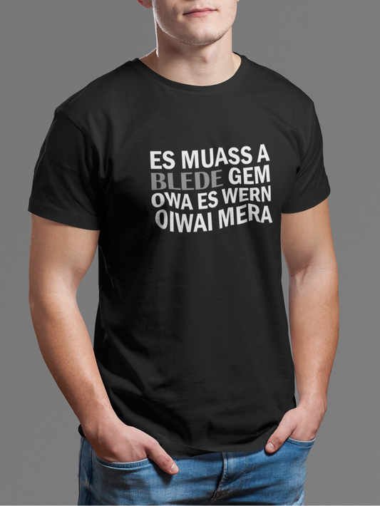 Junger Mann trägt schwarzes T-Shirt mit weißem Text „Es muass a blede gem. Owa es wern oiwai mehr“, Oberkörperansicht