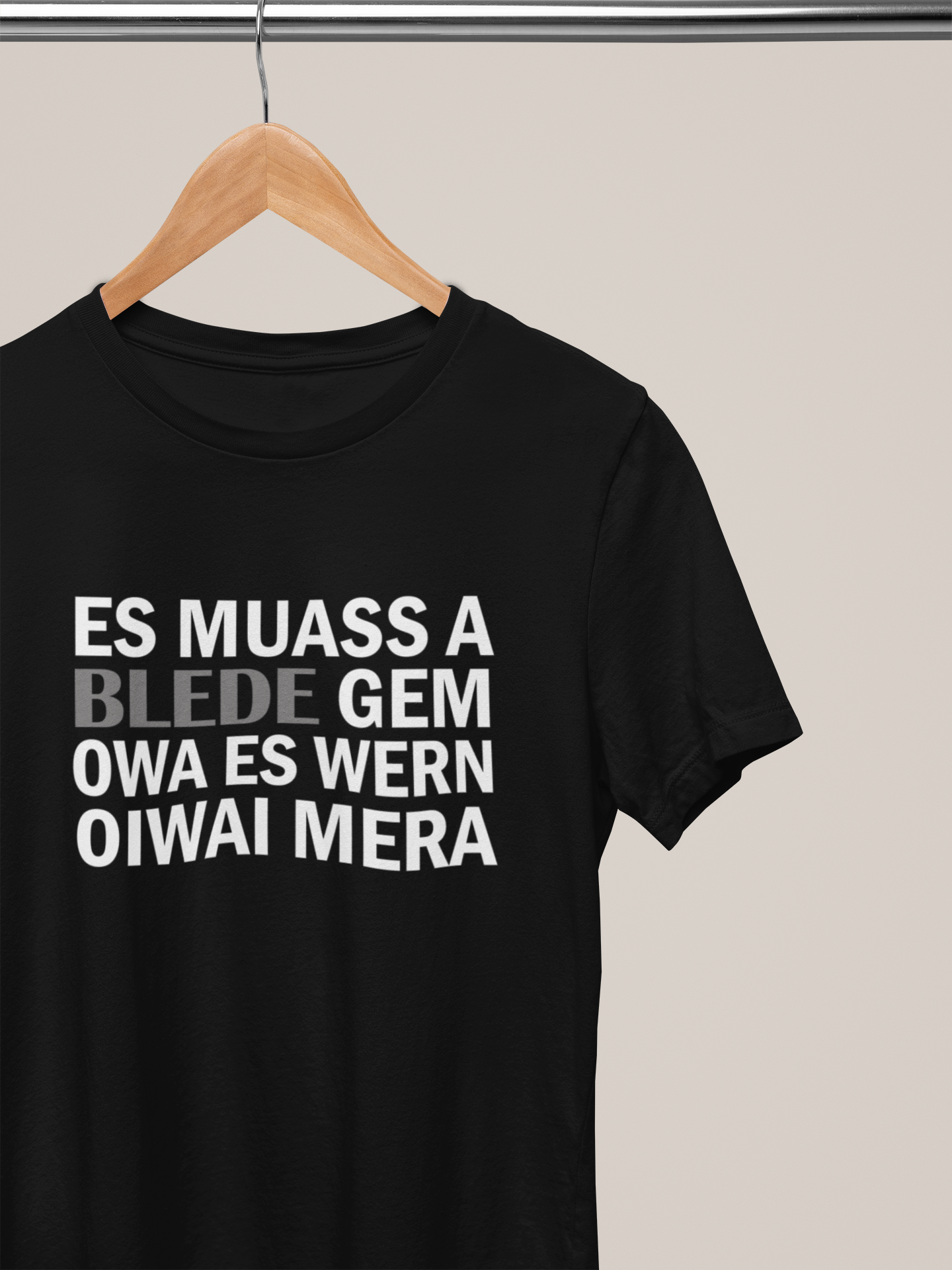 Schwarzes T-Shirt mit weißem Text „Es muass a blede gem. Owa es wern oiwai mehr“, auf Kleiderbügel hängend.