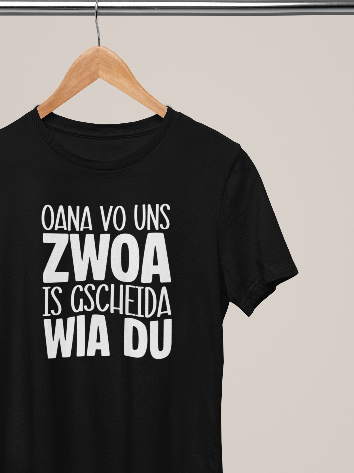 Schwarzes T-Shirt mit weißem Text „Oana vo uns Zwoa is g’scheida wia du“ auf Kleiderbügel 