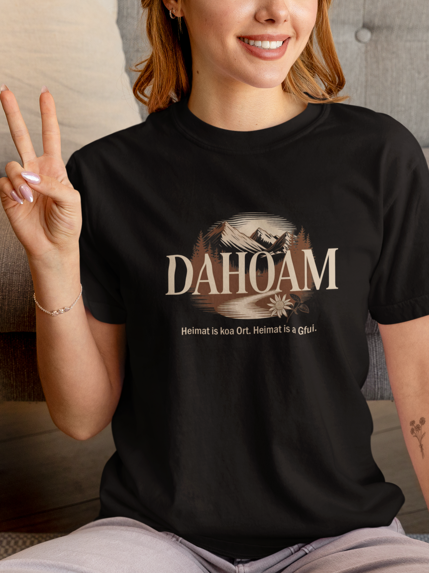 Junge Frau, sitzend, trägt schwarzes T-Shirt mit Bergmotiv in Sepia und Text „Dahoam. Heimat is koa Ort. Heimat is a Gfui.“, Oberkörperansicht