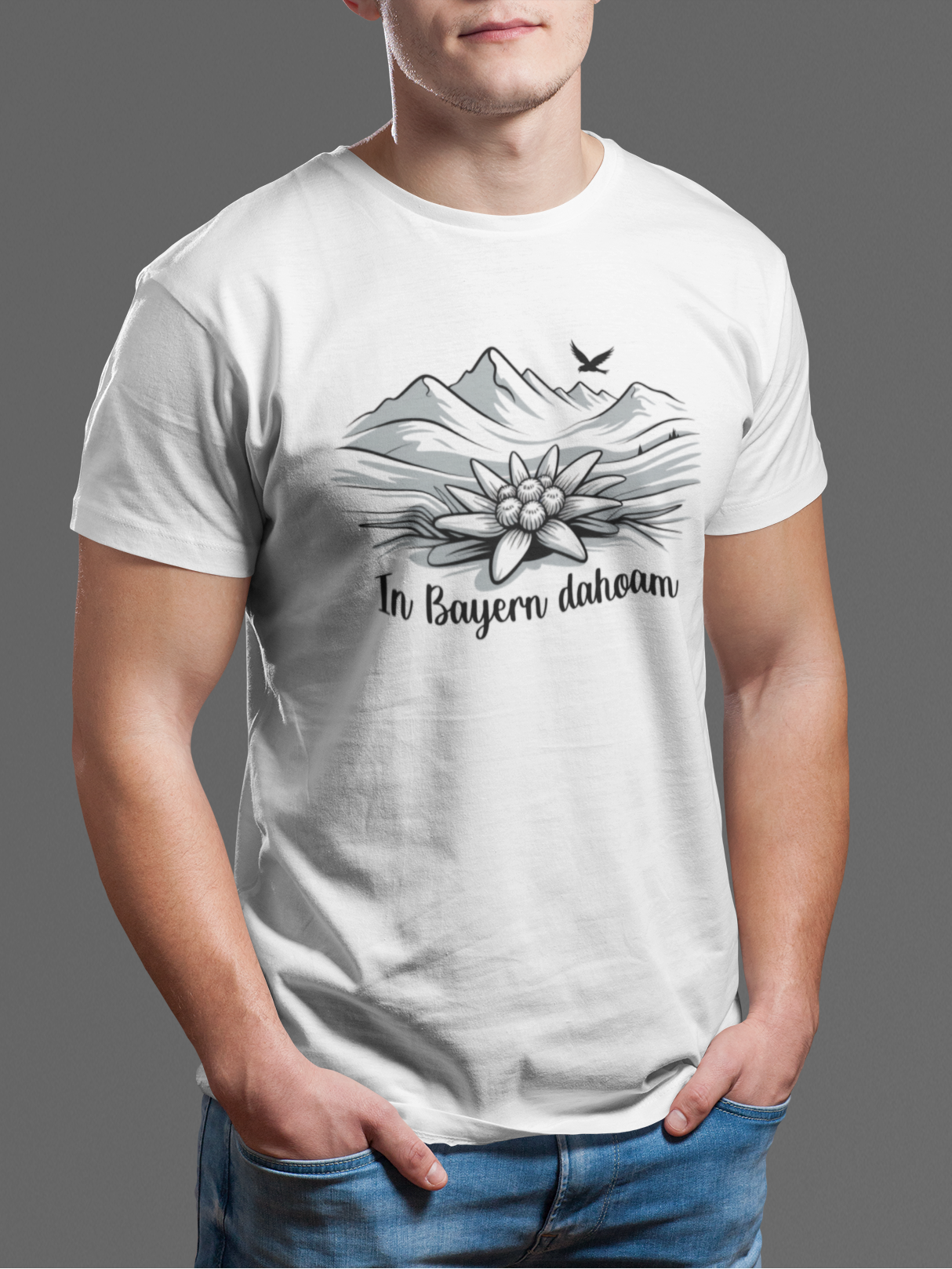 Junger Mann trägt weißes T-Shirt mit Bergpanorama, Edelweiß und Text „In Bayern Dahoam“, Oberkörperansicht