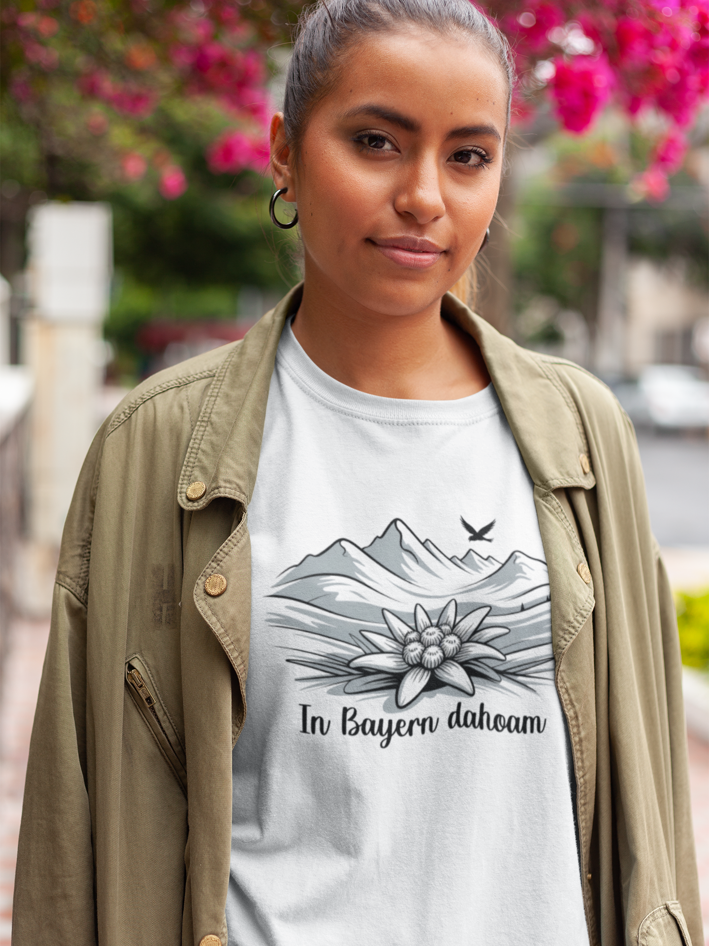 Junge Frau mit beiger offener Jacke, darunter trägt sie ein weißes T-Shirt mit Bergpanorama, Edelweiß und Text „In Bayern Dahoam“, Oberkörperansicht