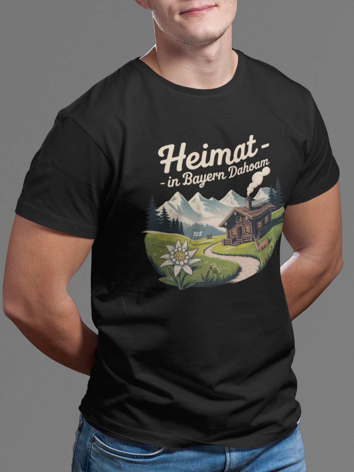 Junger Mann trägt schwarzes T-Shirt mit Motiv „Heimat – In Bayern dahoam“, inklusive Berghintergrund und Almhütte, Oberkörperansicht